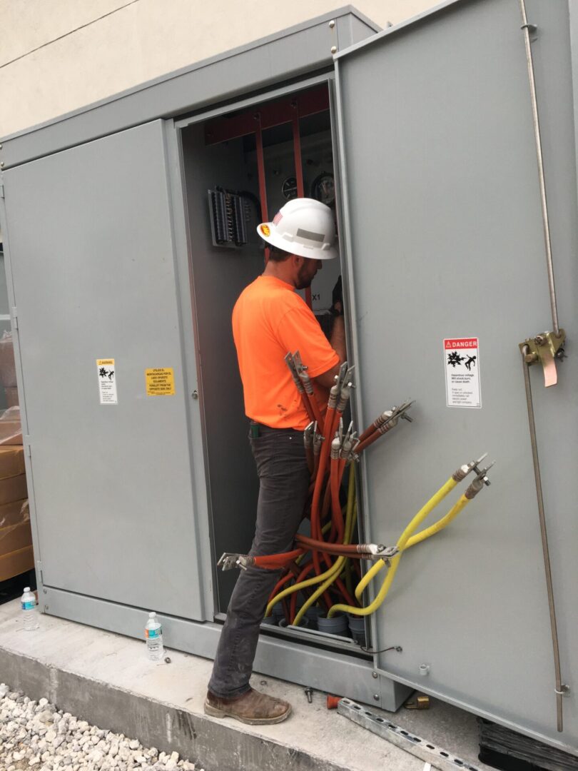 A man in an orange shirt and hard hat working on electrical equipment.